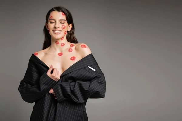 seductive woman with red kiss prints on face and naked shoulders posing in oversize blazer isolated on grey