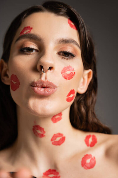 портрет красивой женщины с макияжем и красными отпечатками поцелуев надутых губ при взгляде на камеру, изолированную на сером