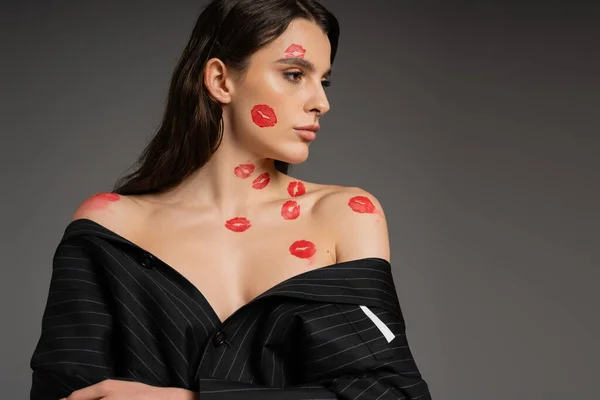 brunette woman with red kiss prints posing in black oversize jacket with bare shoulders isolated on grey