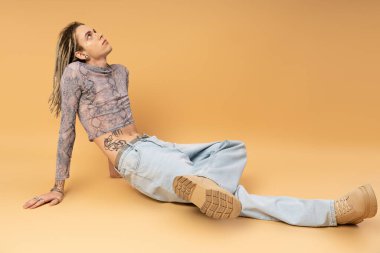 Young queer person in crop top and jeans sitting on yellow background 