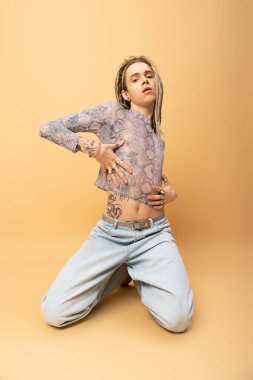 Trendy tattooed queer person touching crop top with snakeskin print on yellow background 