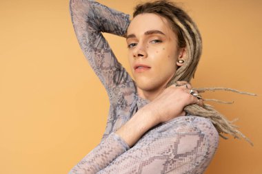 Portrait of tattooed queer person touching dreadlocks and looking at camera isolated on yellow 