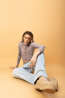 Stylish queer person in jeans and crop top sitting on yellow background 