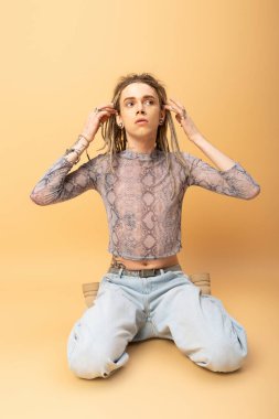 Queer person in crop top with snakeskin print and jeans touching dreadlocks on yellow background 