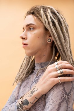 Portrait of tattooed queer person with dreadlocks touching shoulder isolated on yellow 