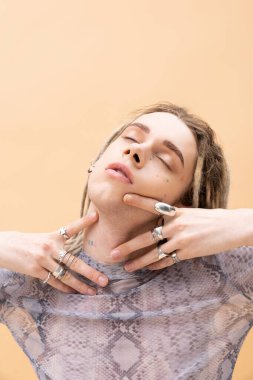 Tattooed nonbinary person with silver accessories touching neck isolated on yellow 
