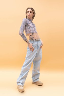 Full length of trendy nonbinary person posing in crop top and jeans on yellow background 