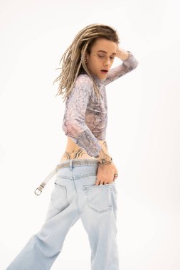 Tattooed queer person in crop top with snakeskin print and jeans posing isolated on white 