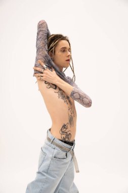 Tattooed queer person in jeans touching crop top isolated on white 