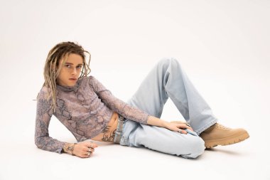 Tattooed nonbinary person in crop top with snakeskin print and jeans lying on white background