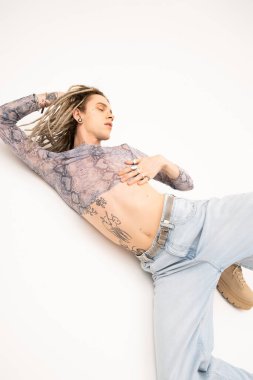 Young tattooed queer person in jeans and crop top posing on white background