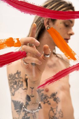 colorful paint strokes on glass near blurred nonbinary person pointing with finger on white background clipart
