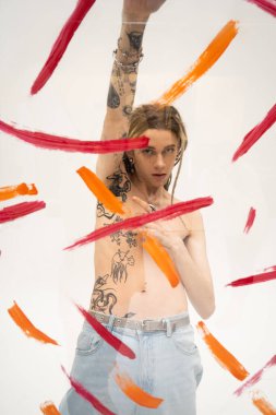 shirtless queer person with tattooed body posing with raised hand behind glass with brush strokes on white background