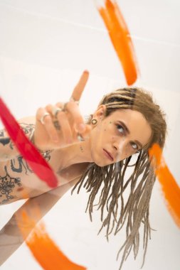 young tattooed queer person with dreadlocks pointing at glass with colorful paint strokes on white background