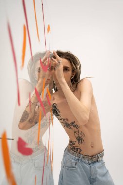 shirtless tattooed queer person in jeans posing near glass with colorful paint on white background