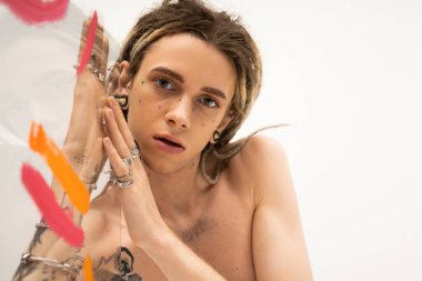 portrait of shirtless queer model with dreadlocks and silver rings looking at camera near glass with colorful paint on white background
