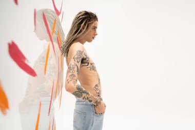 side view of shirtless tattooed nonbinary model posing with hand in pocket of jeans near glass with paint strokes on white background