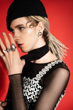 nonbinary model with dreadlocks and silver finger rings holding hands near face and looking away on red 