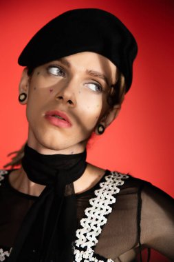 portrait of queer person in black beret and scarf looking away on red background