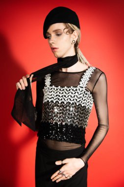 queer person in top with sequins touching black scarf on red background with shadow