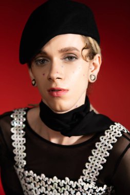 portrait of elegant nonbinary model in black beret and neckerchief looking at camera on red background