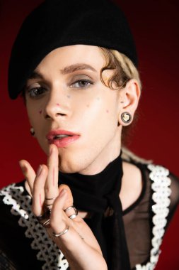 portrait of stylish queer person in makeup and silver rings touching lip and looking at camera on dark red background