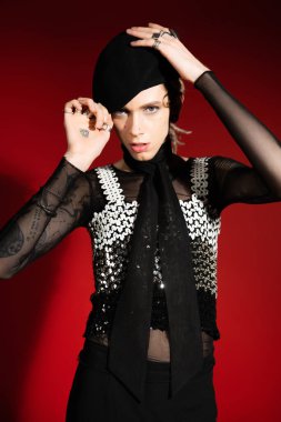 tattooed queer person in top with sequins adjusting black beret and looking at camera on red background