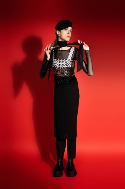 full length of nonbinary model in beret and elegant top tying neckerchief on red background with shadow