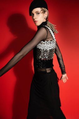 young nonbinary model in elegant top with sequins looking at camera on red with shadow