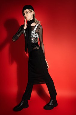 full length of smiling nonbinary person in black elegant clothes holding hand near face on red background with shadow