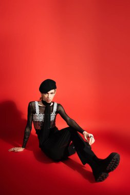 full length of trendy queer model in black beret and boots sitting and looking at camera on red background with shadow