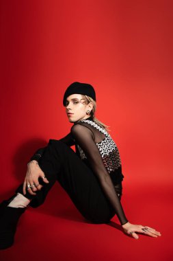 young queer model in black beret and top with sequins sitting and looking at camera on red background