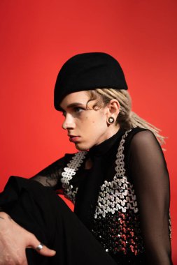 stylish queer person in elegant clothes and black beret looking away while sitting isolated on red