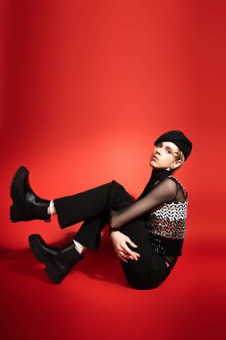 full length of stylish queer model in black leather boots and beret sitting and looking at camera on red background