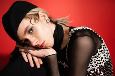 portrait of queer person in black beret and elegant top with sequins looking at camera on red background