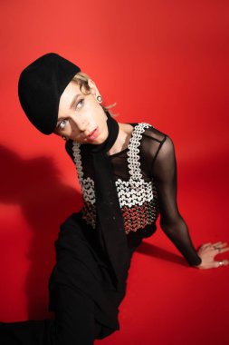 high angle view of trendy nonbinary person in black beret and top with sequins looking away on red background with shadow