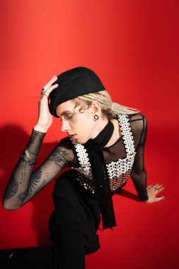 high angle view of elegant nonbinary model touching black beret while sitting with closed eyes on red background