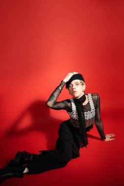 queer person in black and elegant outfit touching beret and looking at camera on red background with shadow