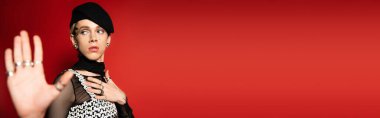 elegant nonbinary person in black beret showing stop gesture isolated on red, banner