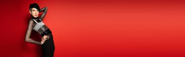 elegant queer model in black beret and elegant top holding hand on hip and looking at camera on red background, banner