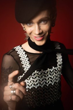 smiling queer model in elegant top and black beret looking at camera and pointing with finger on dark red background