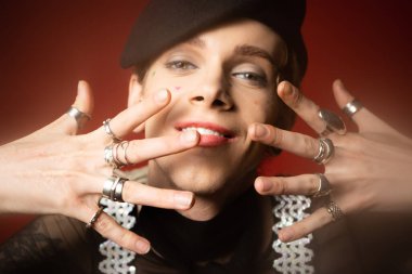smiling queer person in black beret and silver rings posing with hands near face on dark red background