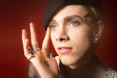 portrait of queer model in black beret and silver rings touching face and looking away on dark red background