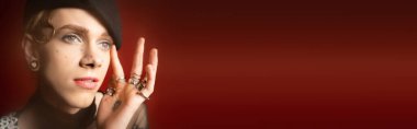 portrait of stylish nonbinary model in silver rings and black beret looking away on dark red background, banner