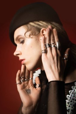 portrait of tattooed nonbinary model in silver rings and black beret on dark red 