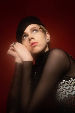 portrait of young queer person in elegant top and black beret looking away on dark red background