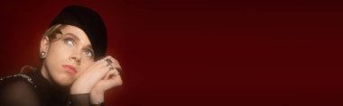 portrait of young queer person in beret holding hands near face and looking away isolated on brown, banner