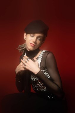 positive queer model in fashionable outfit and silver rings posing with closed eyes and hands near chest on dark red background