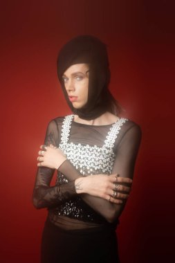 nonbinary person in black headwear and top with sequins posing with crossed arms on dark red background
