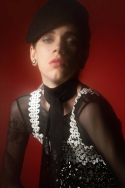 young queer model in black beret and elegant outfit looking at camera on dark red background